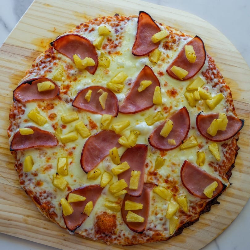 A whole RigaTony's Hawaiian pizza with ham and pineapple on a wooden paddle.