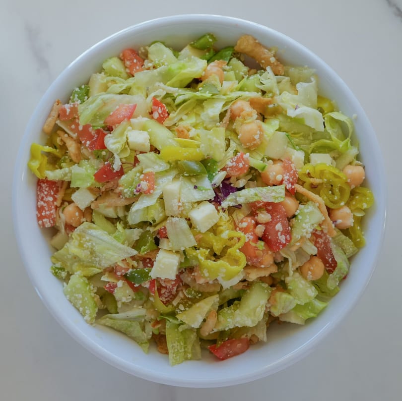 RigaTony’s house salad with lettuce, tomatoes, chickpeas, cheese, and pepperoncini in a white bowl.
