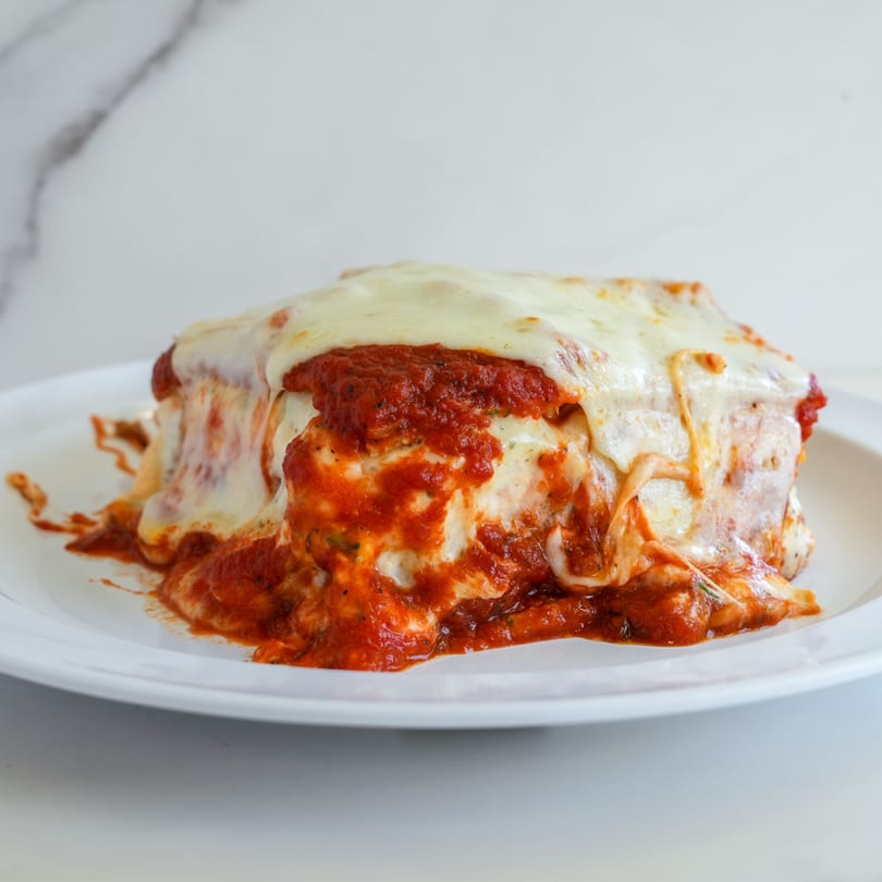 A generous serving of RigaTony’s lasagna, topped with melted cheese and rich red sauce, on a white plate.