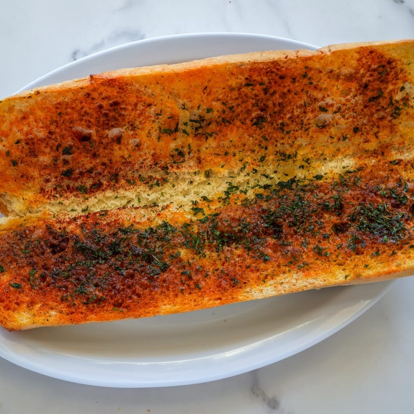 A close-up of RigaTony's garlic bread, golden brown with herbs and spices, on a white plate.