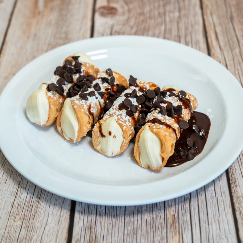 A plate of RigaTony's cannolis, drizzled with chocolate sauce, chocolate chips, and powdered sugar.