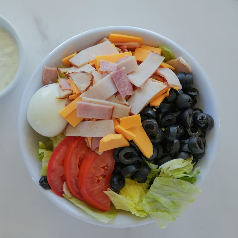 A white bowl filled with a salad featuring lettuce, tomatoes, olives, cheese, ham, turkey, and a hard-boiled egg.