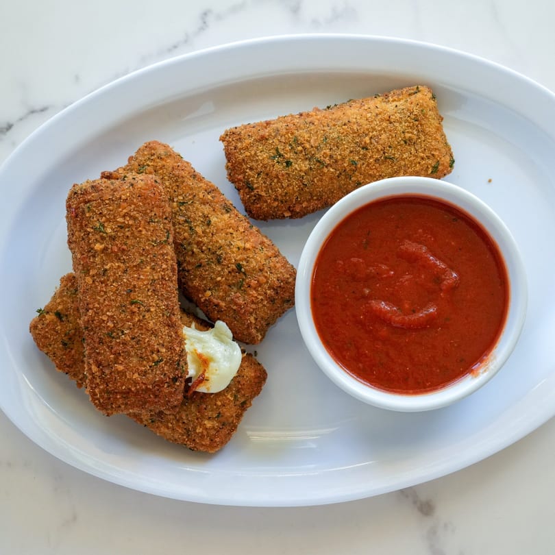 Golden-brown mozzarella sticks with melted cheese and a side of red marinara sauce on a white plate.