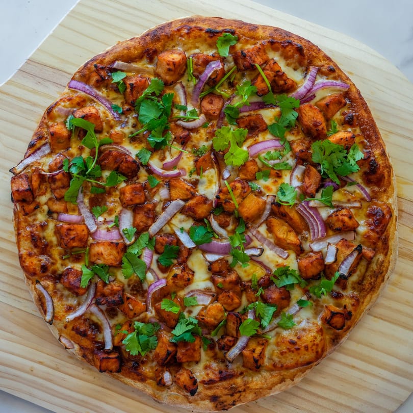 A RigaTony's pizza topped with chicken, red onions, and cilantro on a wooden board.