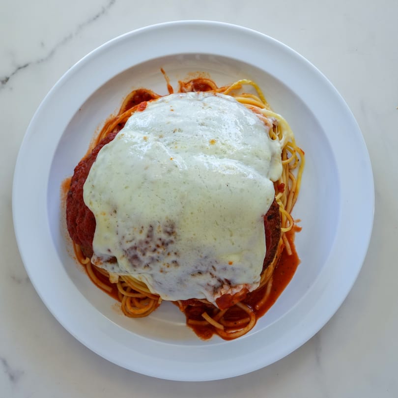 A white plate holds spaghetti with marinara sauce, topped with a large chicken parmesan cutlet covered in melted cheese.