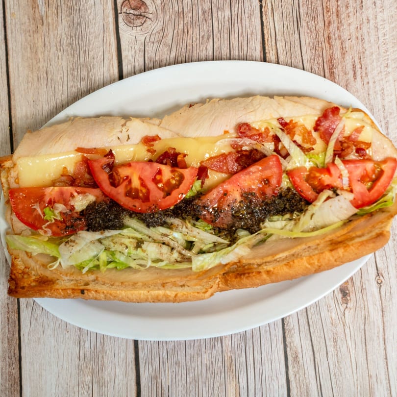 A RigaTony’s sub sandwich with turkey, bacon, cheese, tomatoes, lettuce, and pesto on a white plate.